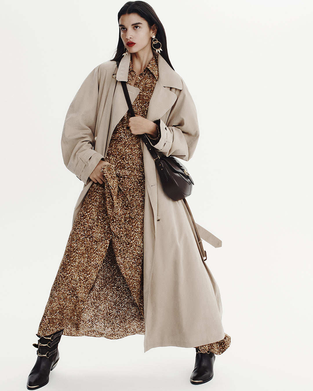 Model wears a long printed dress in brown and yellow tones with a trench and boots with metallic details.
