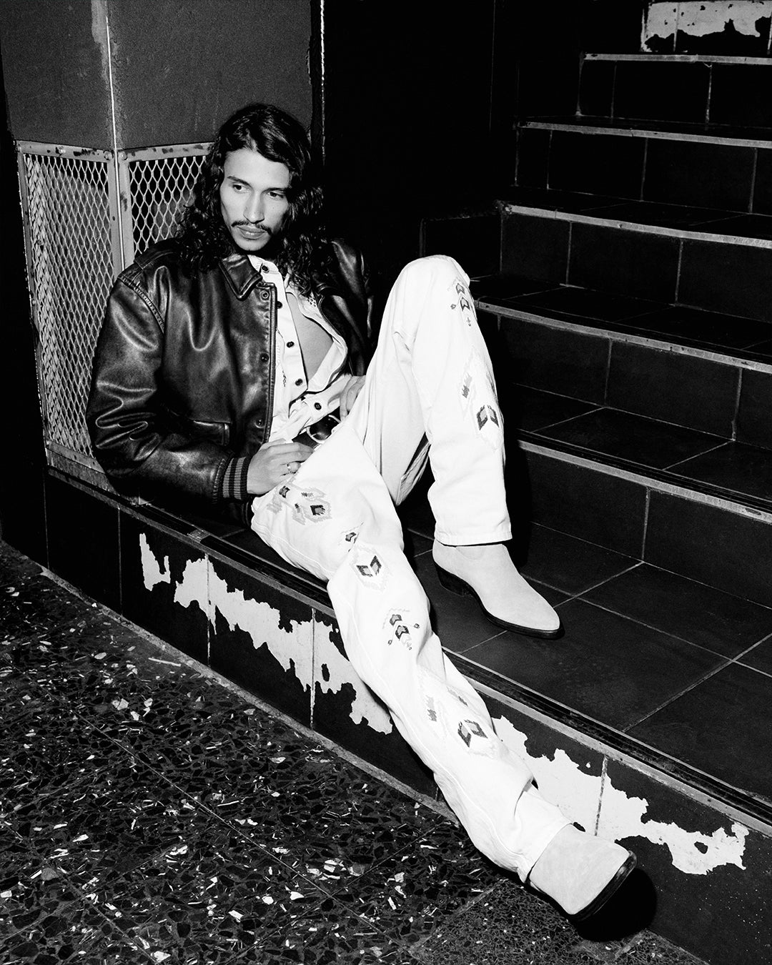 Men seats in the stairs wearing a leather jacket and a white embroidered pants.