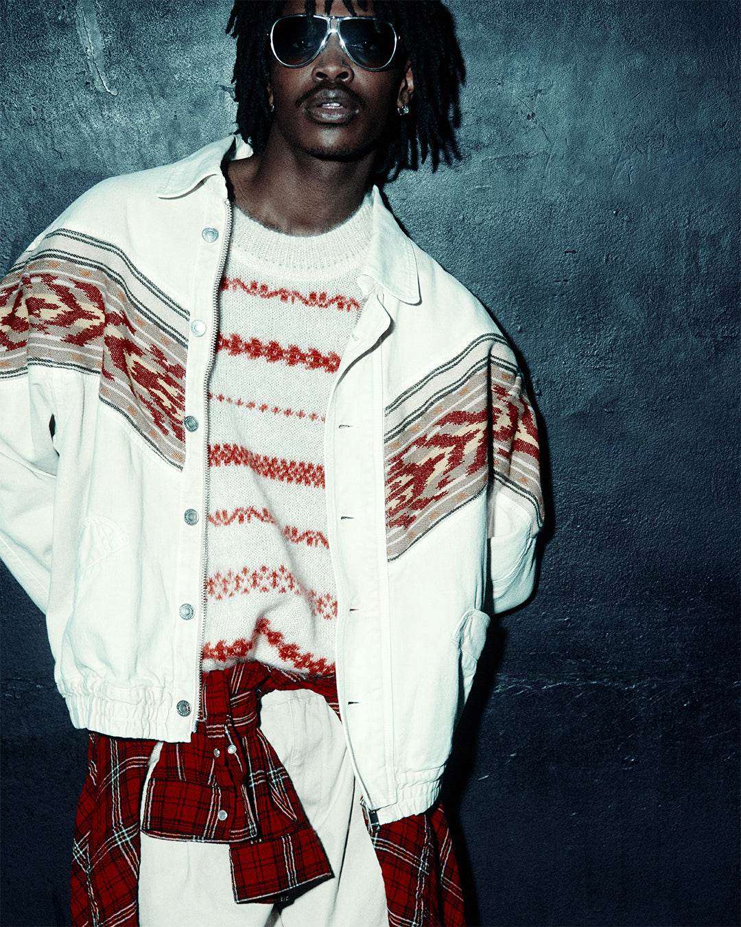 Male model in folk-inspired open shirt with red-black stripes, bare chest, paired with black leather trousers, photographed in a dimly lit club setting.