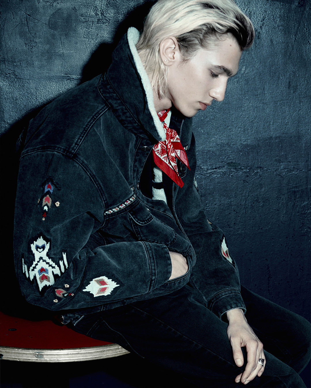 Embroidered denim jacket with artisanal detailing worn over casual pants, model in relaxed pose in underground-chic environment.