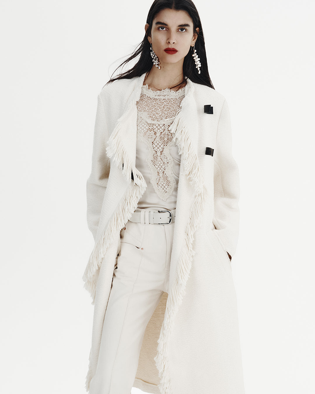 Paola wears long coat and lace top with a pant and belt, all white.