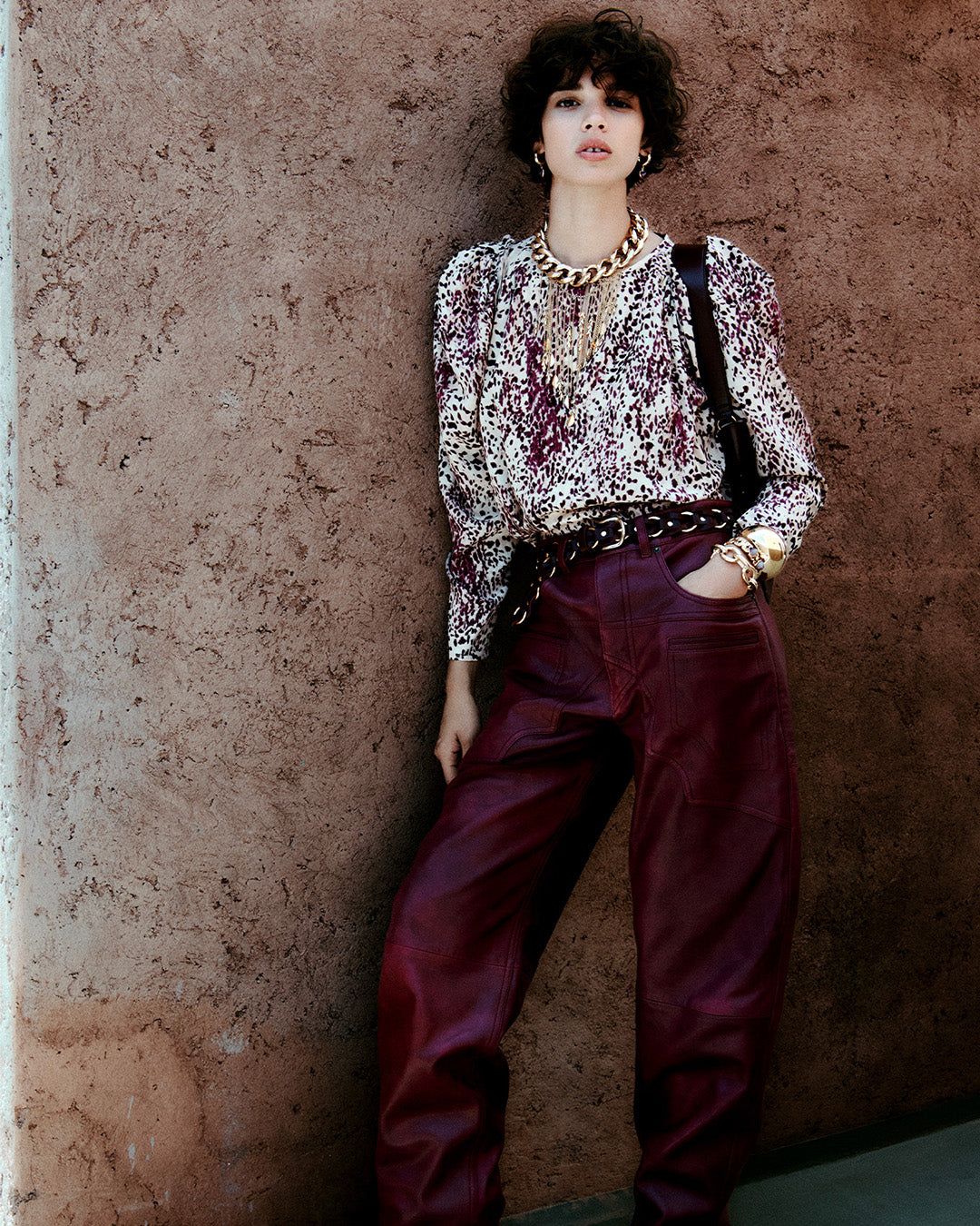 Feminine model, dressed in the Zarga blouse in printed silk with purple and ecru pleats, the Breydi pants in lambskin with berry-colored cut-outs and the Lyka santiag boots in leather with straps.