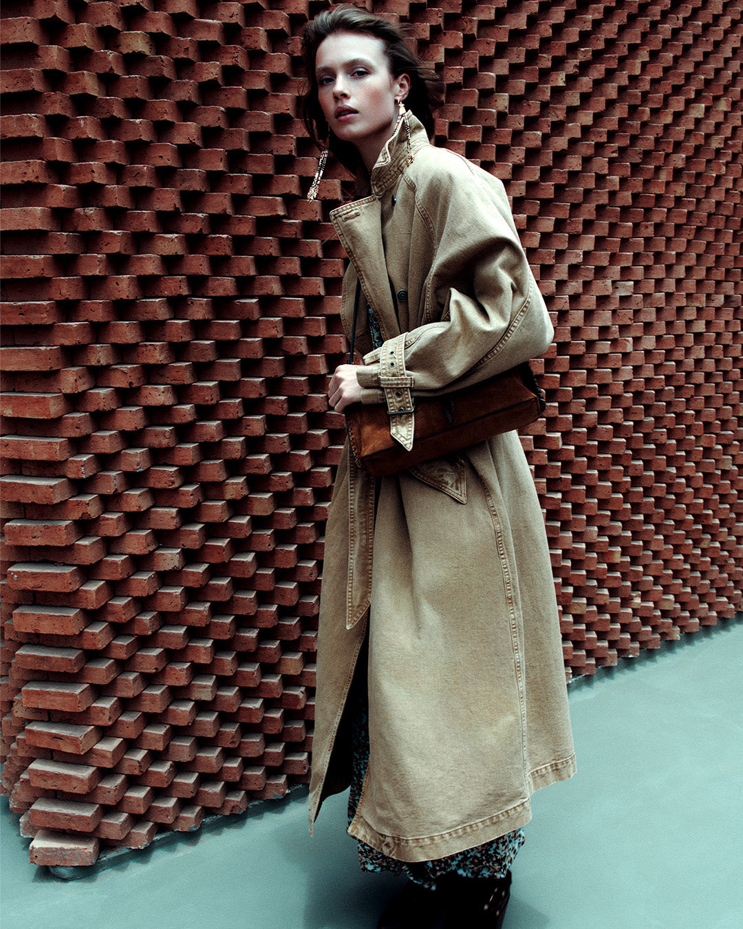 A feminine model, dressed in a long camel trench coat with Lyka santiag boots in leather with black straps.