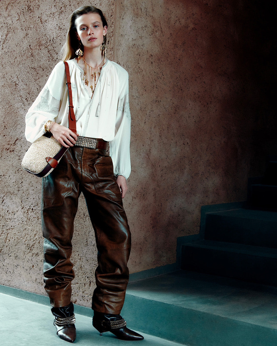 Feminine model, dressed in the Oris blouse in ecru embroidered crepe georgette, loose-fitting pants, Edrik draped boots with black chains and the Bayia bucket bag in raffia.