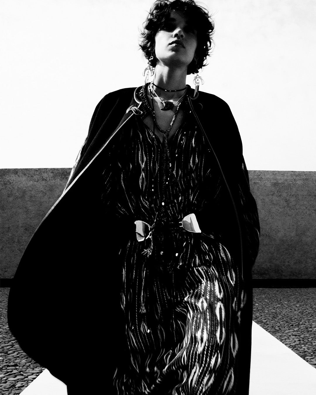 Feminine model, dressed in a long cape in wool and cashmere and the Sana long dress in silk with black and ecru graphic motifs