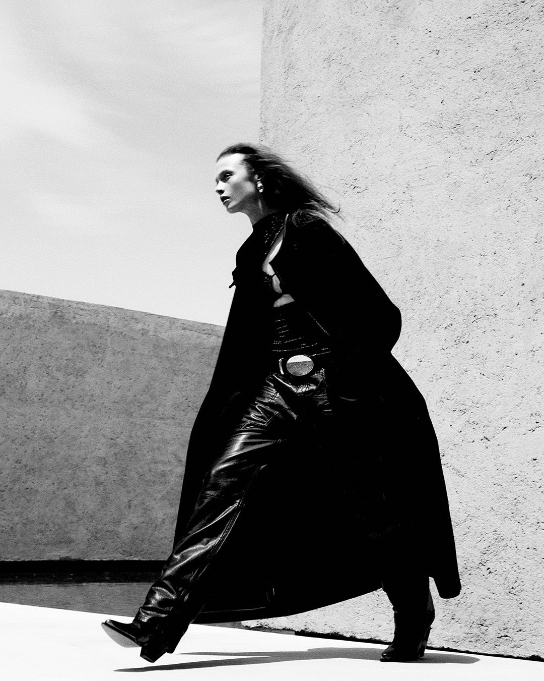 Feminine model, dressed in the classic Elza coat in black wool and cashmere, Breydi pants in lambskin with black cut-outs and Lyka santiag boots in leather with black straps.