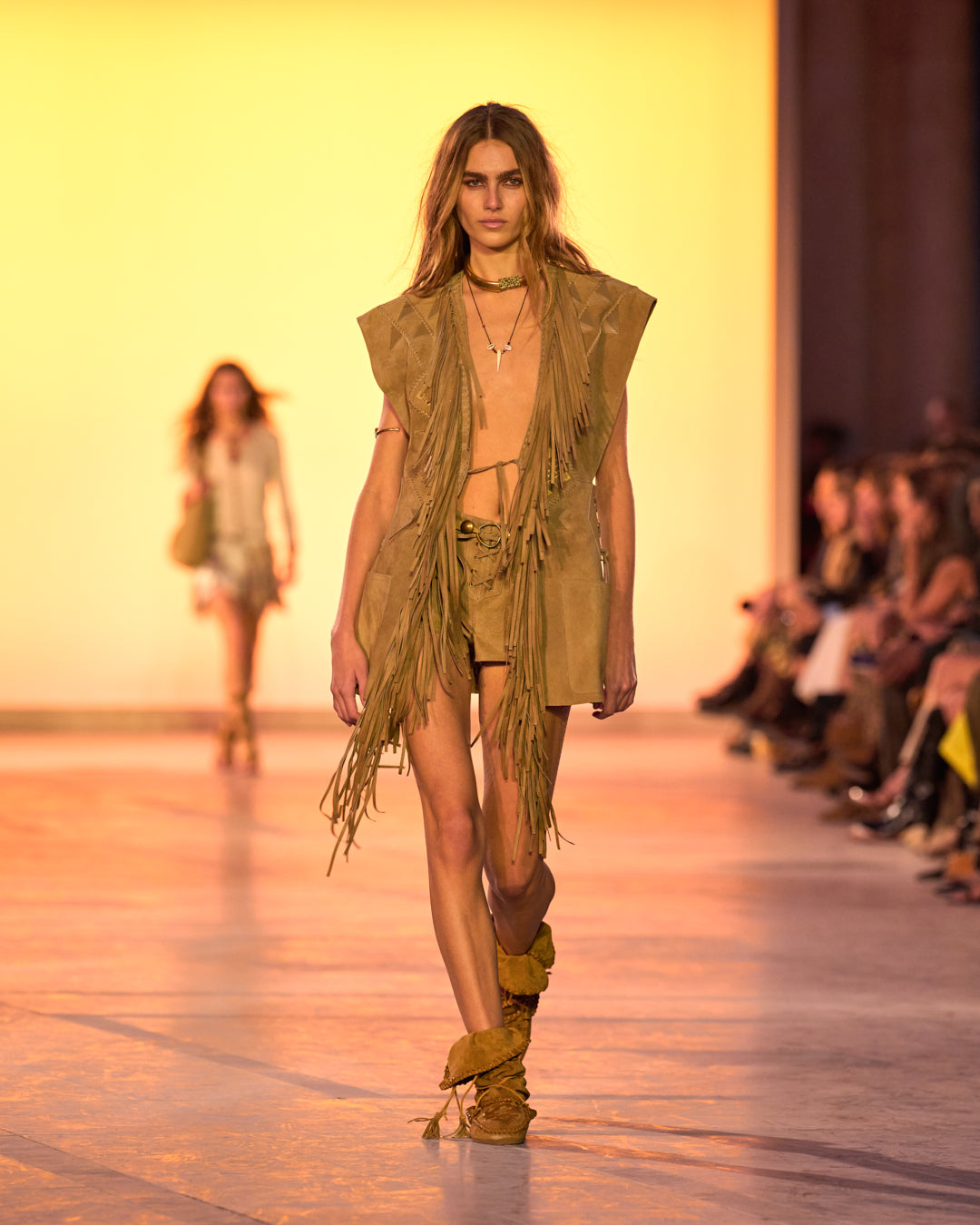 Female model on the catwalk, wearing a Dalila sleeveless cardigan in suede with bangs, shorts and Winki boots in suede and braided leather.