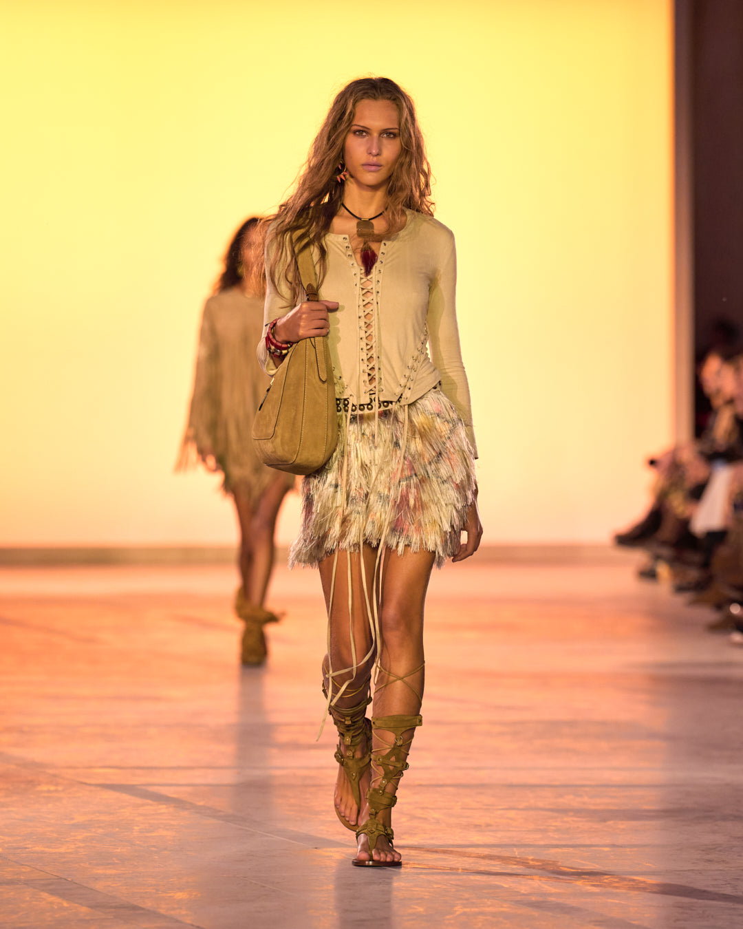 Women's model on the catwalk, wearing Seatle faux suede lace-up top, fringed skirt, Altay Hobo bag and Nensi leather high-top sandals.