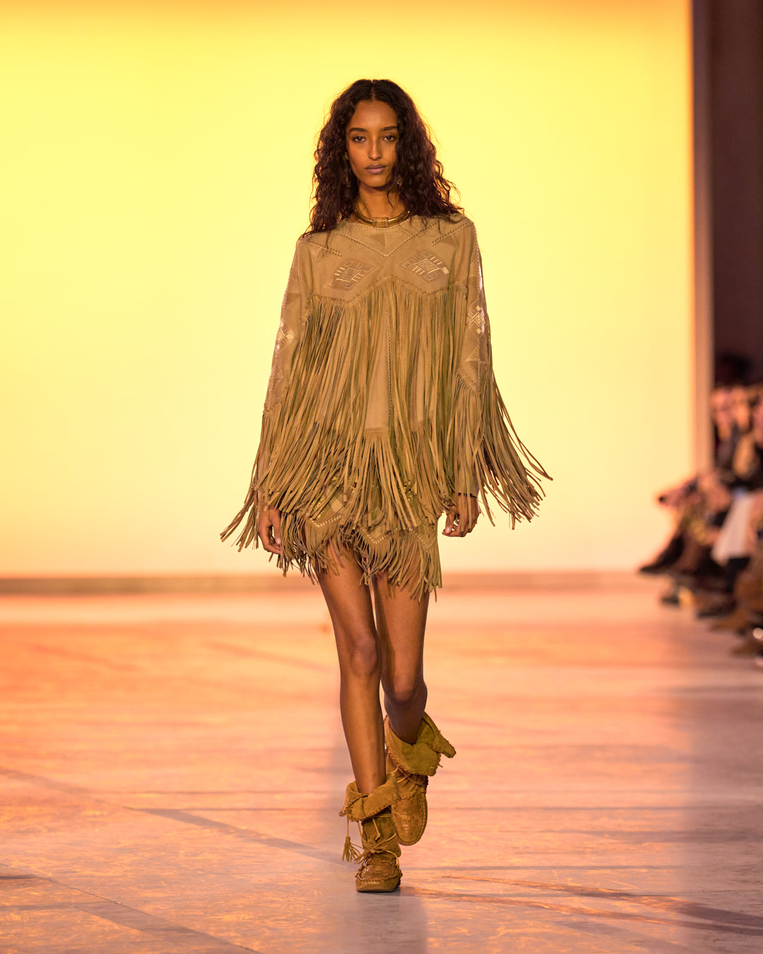 Female model on the catwalk, dressed in a long-sleeved jacket with bangs, a camel skirt with bangs and Winki boots in suede and braided leather.