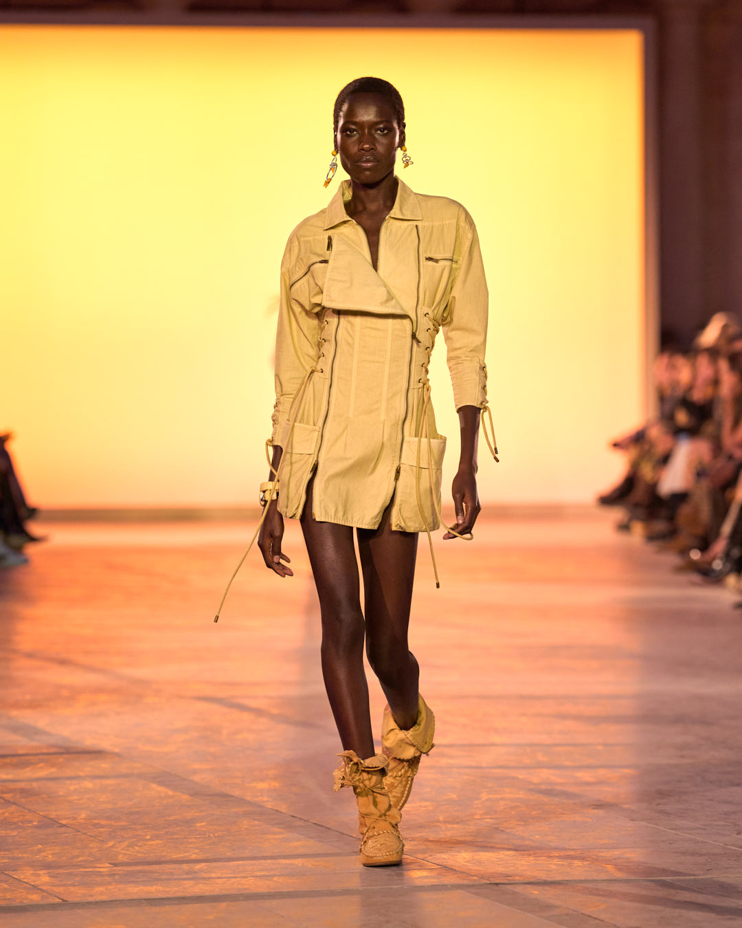Female model on the catwalk, wearing the Izarra short dress with ecru lacing and Winki boots in suede and cognac-coloured braided leather.