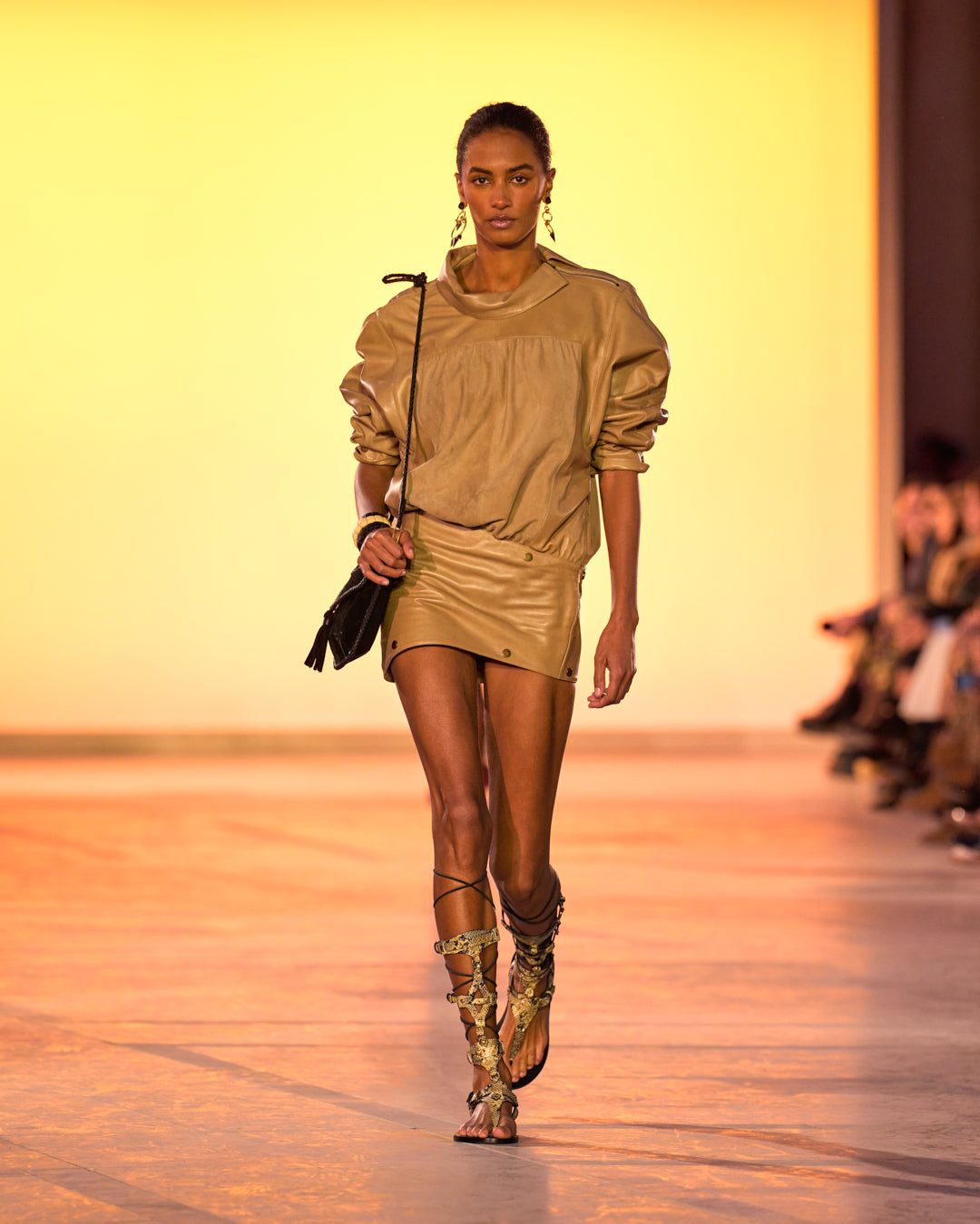 Female model on the catwalk, dressed in a calm faux-leather dress, black Anahi bag and python-print sandals.