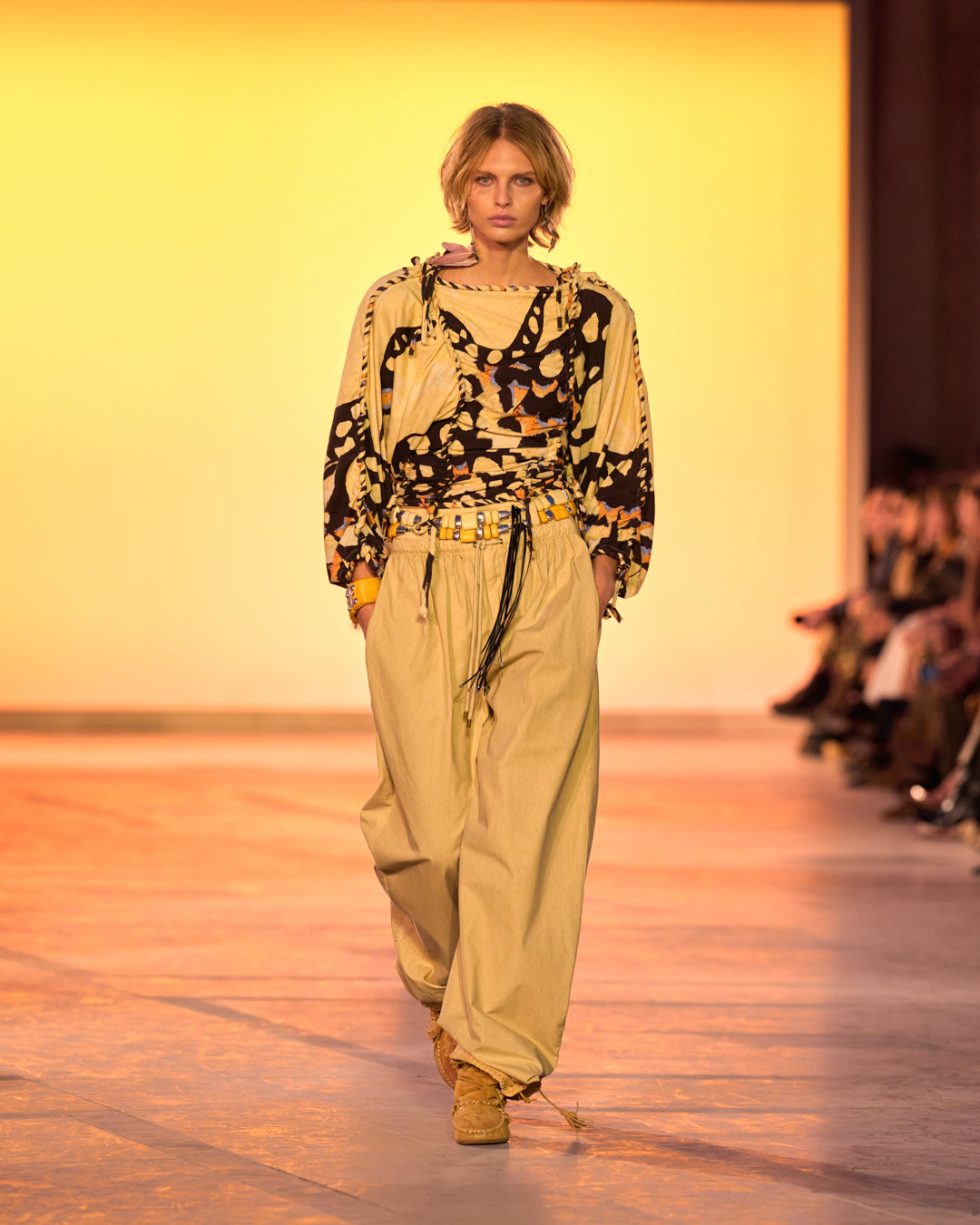 Feminine model on the catwalk, dressed in a printed and braided jersey top, fluid beige pants, the Rosly beaded jewel belt and Winki boots in suede and braided leather in cognac.