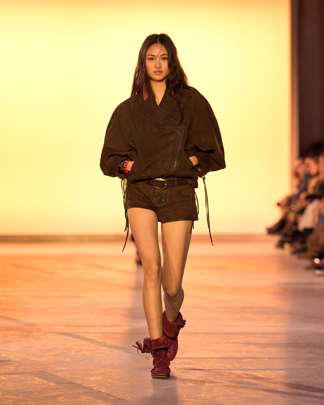 Female model on the catwalk, dressed in an embroidered shirt, the Edina short jacket in suede, dark brown shorts, and Winki boots in suede and braided leather in burgundy.