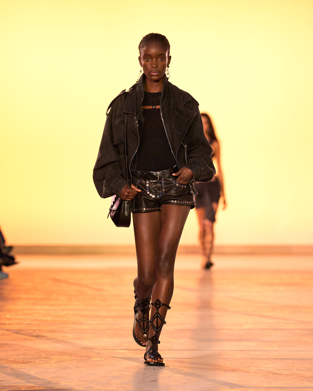 Female model on the catwalk, wearing the Elsie Sweater in fine knit and black pearls, the Elsanna Lace-up Shorts in black studded leather, the Aneli Pocket Jacket in black denim, the altay small bag in black and black sandals.