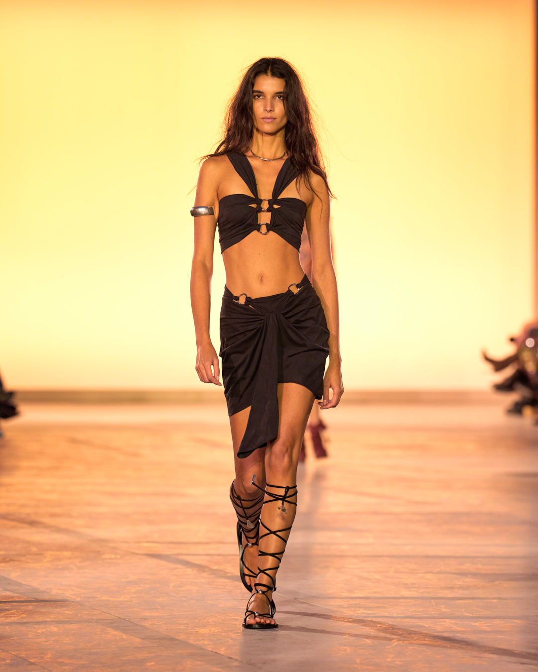 Female model on the catwalk, wearing a black dress and black suede Azzi sandals.