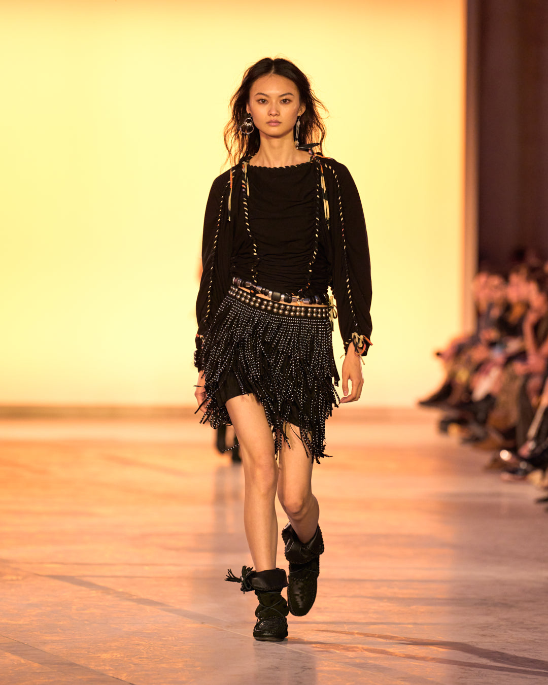 Female model on the catwalk, wearing the Zohra top in black woven jersey, the short Carmelia skirt with black bangs and studs and the Winki boots in black suede and woven leather.