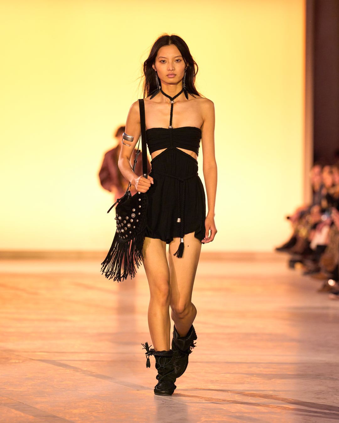 Female model on the catwalk, dressed in a short black dress, Oskan Soft Zipped bag in leather with studs and bangs and Winki boots in black suede and braided leather.