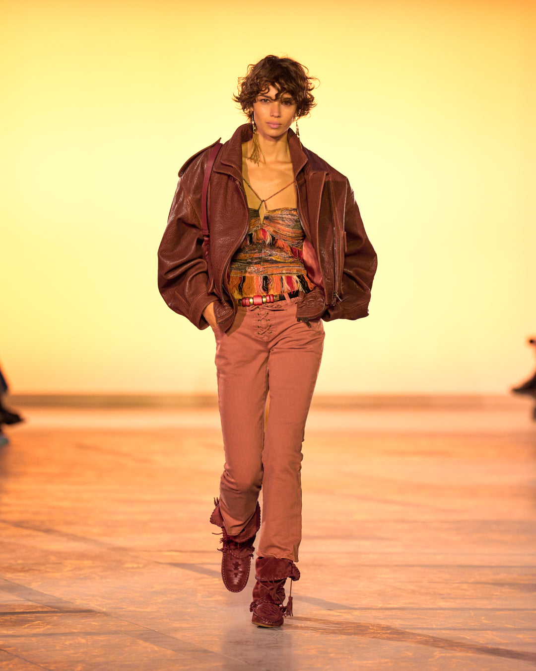 Female model on the catwalk, dressed in a burgundy pocket vest, Darlene strapless top in printed tulle, Vanel lace-up jeans in henna-colored denim and Winki boots in burgundy suede and braided leather.