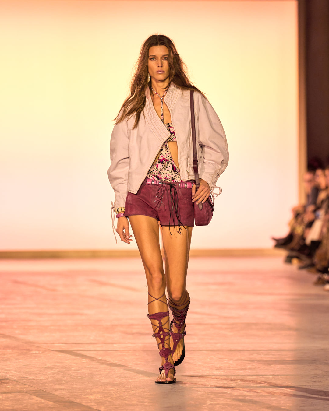 Female model on the catwalk, dressed in a pink denim cropped jacket, altay small in burgundy, Delma lace-up shorts in raspberry suede and raspberry sandals.