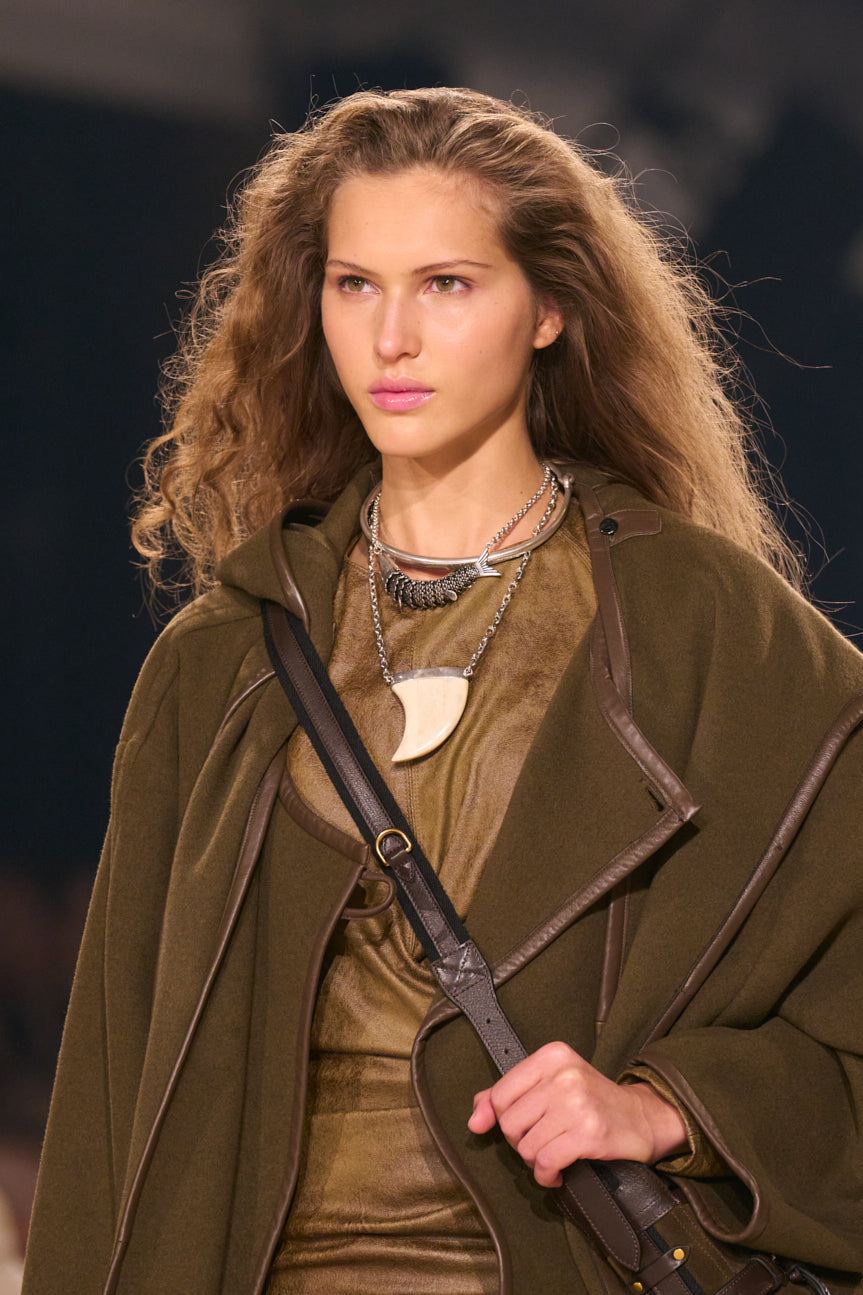 Cropped runway shot of female model wearing a light brown ensemble topped with an oversized khaki-colored coat, carrying a brown cross-body messenger bag.